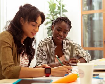 Zwei Frauen arbeiten gemeinsam an einem Tisch, eine schreibt, während die andere aufmerksam zuschaut. Schreibmaterialien und Getränke sind sichtbar.