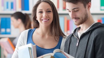 Zwei junge Erwachsene in einer Bibliothek, einer hält Bücher, während die andere lächelt und einen offenen Buchdeckel zeigt.