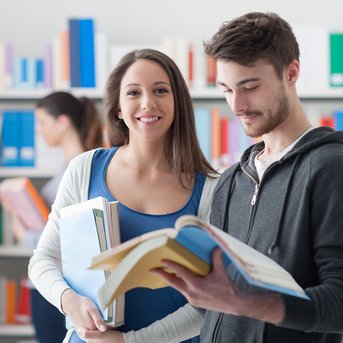 Eine Frau lächelt, während sie mehrere Bücher in den Händen hält. Ein Mann steht neben ihr und schaut auf ein aufgeschlagenes Buch. Im Hintergrund sind Bücherregale mit bunten Büchern sichtbar.