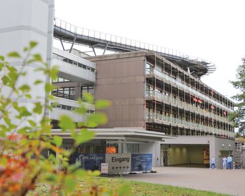 Eingang zu einem modernen Gebäude mit mehreren Etagen, umgeben von Bäumen und Pflanzen. Auf dem Dach verläuft eine Rutsche. Personen in medizinischer Kleidung sind im Eingangsbereich sichtbar.