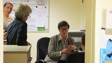 Drei Frauen und ein Mann in einem Büro, die an einem Tisch sitzen und miteinander sprechen. Ein Kalender und Dokumente sind im Hintergrund sichtbar.