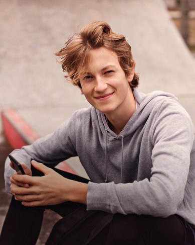 Ein junger Mann mit blonden Haaren sitzt auf dem Boden eines Skateparks, hält ein Smartphone in der Hand und lächelt in die Kamera. Er trägt einen grauen Hoodie und schwarze Hosen.