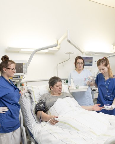 Eine Patientin liegt im Bett in einem Krankenhauszimmer, umgeben von drei Pflegekräften. Eine Pflegekraft misst den Blutdruck der Patientin, während die anderen beiden Informationen bereitstellen und Medikamente vorbereiten. Das Zimmer ist hell und modern eingerichtet.