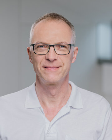 Ein Mann mit Brille und kurzem, grauem Haar trägt ein weißes Polohemd. Er steht in einem modernen, hellen Raum mit großen Fenstern im Hintergrund.