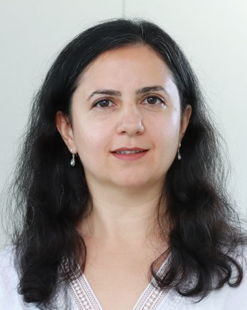 Eine Frau mit langen, lockigen, dunklen Haaren und Ohrringen, die in die Kamera schaut. Sie trägt ein helles Oberteil.