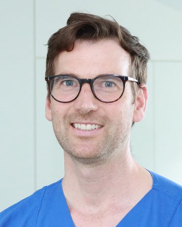 Ein Mann mit Brille und blauer Arbeitskleidung lächelt in die Kamera. Er hat kurze, braune Haare und steht vor einem hellen Hintergrund.