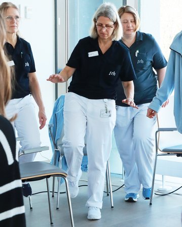 Drei Frauen in blauen Poloshirts und weißen Hosen führen eine Gehübung in einem hellen Raum durch.