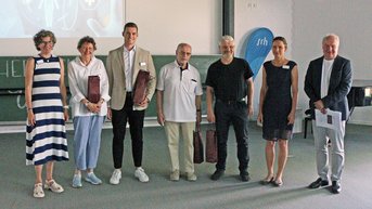 Sechs Personen stehen in einer Reihe, alle in formeller Kleidung, mit Geschenktüten in den Händen. Im Hintergrund ist eine Präsentation sichtbar.