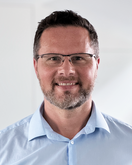 Ein Mann mit Brille und einem kurzen Bart lächelt in die Kamera. Er trägt ein hellblaues Hemd und steht vor einem hellen, neutralen Hintergrund.