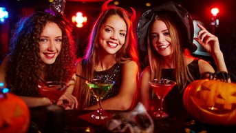 Drei Frauen in Halloween-Kostümen sitzen an einem Tisch mit bunten Cocktails. Sie lächeln und posieren, während Kürbisse und Dekorationen im Hintergrund sichtbar sind. Die Atmosphäre ist festlich und thematisch passend.