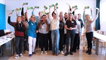 Eine Gruppe von Menschen steht in einem Raum und hebt Karten in die Höhe. Sie lächeln und scheinen an einer Veranstaltung teilzunehmen.