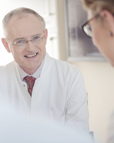 Ein Arzt in einem weißen Kittel lächelt und spricht mit einer Patientin oder Kollegin, die ihm gegenüber sitzt. Die Umgebung ist hell und freundlich.