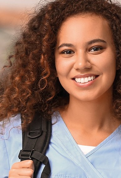 Junge Frau mit lockigem Haar trägt eine hellblaue Uniform und einen Rucksack. Sie lächelt freundlich in die Kamera. Im Hintergrund sind weitere Personen in ähnlicher Kleidung zu sehen.