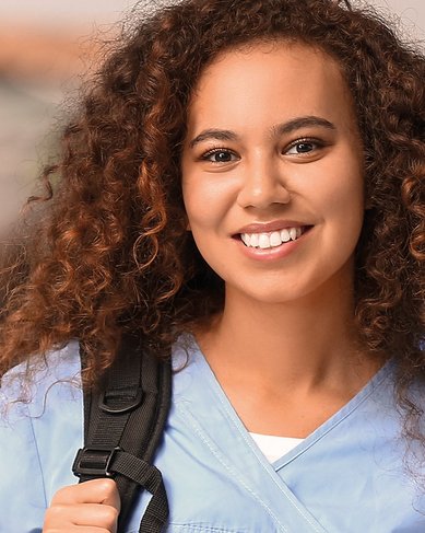 Junge Frau mit lockigem Haar trägt eine hellblaue Uniform und einen Rucksack. Sie lächelt freundlich in die Kamera. Im Hintergrund sind weitere Personen in ähnlicher Kleidung zu sehen.
