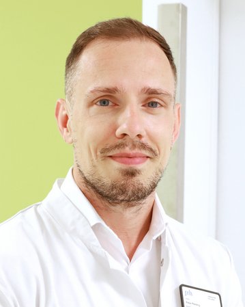 Ein Mann mit kurzen, hellen Haaren und einem gepflegten Bart trägt einen weißen Kittel. Er lächelt und steht vor einer grünen Wand.