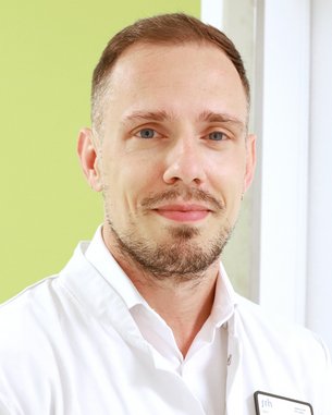 Ein Mann mit kurzen, hellen Haaren und einem gepflegten Bart trägt einen weißen Kittel. Er lächelt und steht vor einer grünen Wand.