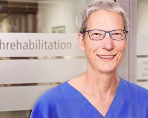 Eine lächelnde Frau in blauer Arbeitskleidung steht vor einem Fenster mit der Aufschrift „Frührehabilitation“.