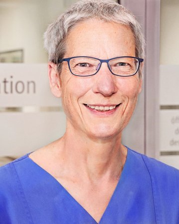 Eine lächelnde Frau in blauer Arbeitskleidung steht vor einem Fenster mit der Aufschrift „Frührehabilitation“.