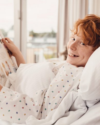 Ein lächelndes Kind mit roten Haaren liegt im Bett und hält ein Buch in der Hand. Es trägt ein gepunktetes Nachthemd und schaut in die Kamera. Im Hintergrund sind Fenster mit Blick auf eine Stadtlandschaft zu sehen.