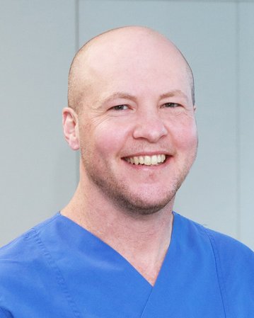 Ein Mann mit Glatze trägt ein blaues medizinisches Outfit und lächelt freundlich in einem hellen Raum.