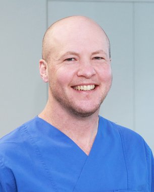 Ein Mann mit Glatze trägt ein blaues medizinisches Outfit und lächelt freundlich in einem hellen Raum.