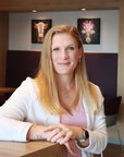 Eine Frau mit langen, blonden Haaren sitzt an einem Tisch in einem modernen Raum. Sie trägt ein weißes Oberteil und lächelt leicht. Im Hintergrund sind Pflanzen und Kunstwerke an der Wand zu sehen.