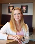 Eine Frau mit langen, blonden Haaren sitzt an einem Tisch in einem modernen Raum, lächelnd und mit einer Pflanze im Hintergrund.