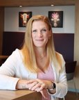 Eine Frau mit langen, blonden Haaren sitzt an einem Tisch in einem modernen Raum, lächelnd und mit einem Pflanzenarrangement im Hintergrund.
