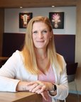 Eine Frau mit langen, blonden Haaren sitzt an einem Tisch in einem modernen Raum, lächelnd und mit einem Pflanzenarrangement im Hintergrund.