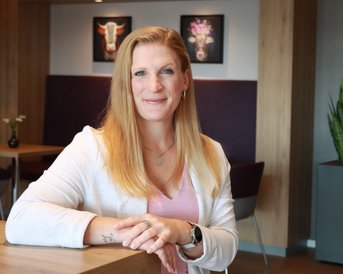Eine Frau mit langen, blonden Haaren sitzt an einem Tisch in einem modernen Raum. Sie trägt ein weißes Oberteil und lächelt leicht. Im Hintergrund sind Pflanzen und Kunstwerke an der Wand zu sehen.