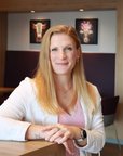 Eine Frau mit langen, blonden Haaren sitzt an einem Tisch in einem modernen Raum, lächelt und trägt einen weißen Cardigan.