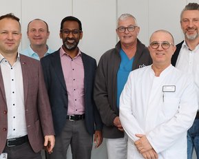 Sechs Männer stehen in einer Reihe, lächelnd, in formeller und medizinischer Kleidung, vor einer neutralen Wand.