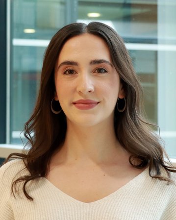 Eine junge Frau mit langen, lockigen Haaren und Ohrringen, die in einem hellen Pullover vor einem modernen Hintergrund steht.