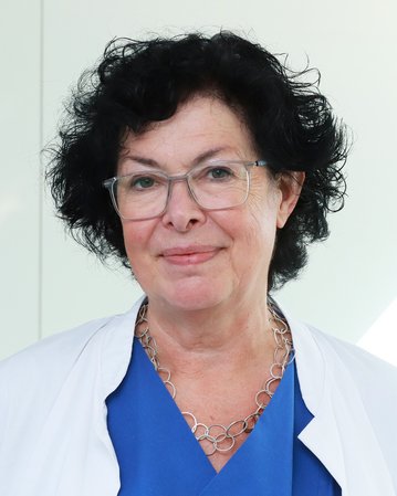 Ältere Frau mit lockigem, schwarzem Haar, Brille und medizinischer Kleidung, lächelt freundlich in einem hellen Raum.