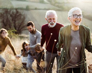 Eine Gruppe von fünf Personen wandert fröhlich einen Hügel hinauf, während eine ältere Frau mit Brille und einem Korb in der Hand an der Spitze steht. Die anderen folgen ihr, lächelnd und in lockerer Kleidung.