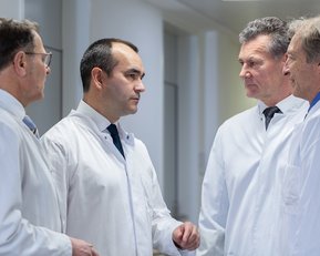 Vier Ärzte in weißen Kitteln stehen in einem Flur und diskutieren miteinander. Im Hintergrund ist ein Blumenbild zu sehen.