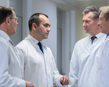 vlnr: Prof. Tobias Pitzen, Prof. Timo Zippelius, Dr. Gregor Ostrowski und PD Dr. Michael Ruf stehen in einem Flur und diskutieren miteinander. Im Hintergrund ist ein Bild mit Blumen zu sehen.