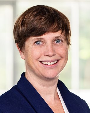 Eine Person mit kurzen, braunen Haaren und einem freundlichen Lächeln trägt einen blauen Blazer. Der Hintergrund zeigt unscharfe Fenster und eine helle, neutrale Umgebung.