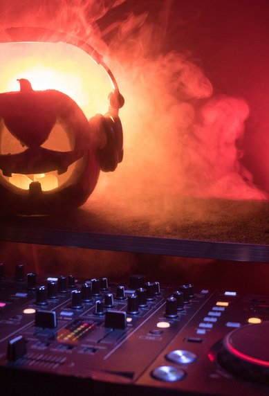 Ein leuchtender Kürbis mit Kopfhörern steht auf einem DJ-Pult, umgeben von Nebel und bunten Lichtern. Die Kürbislaterne hat ein gruseliges Gesicht und schafft eine festliche Halloween-Atmosphäre.