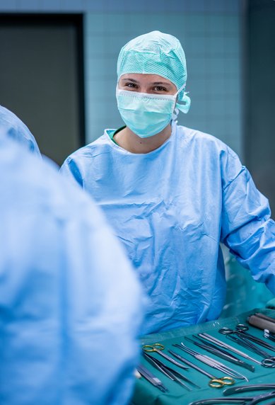 Eine Person in blauer OP-Kleidung und Mundschutz steht in einem Operationssaal, umgeben von chirurgischen Instrumenten.