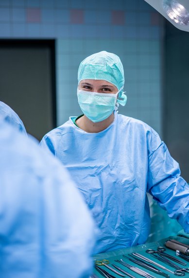 Eine Person in blauer OP-Kleidung und Mundschutz steht in einem Operationssaal, umgeben von chirurgischen Instrumenten.