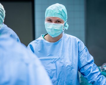 Eine Person in blauer OP-Kleidung und Mundschutz steht in einem Operationssaal, umgeben von chirurgischen Instrumenten.