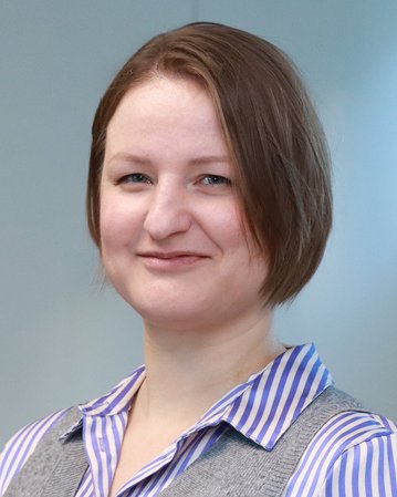 Eine Person mit kurzen, braunen Haaren und einem grauen Pullover mit blauer gestreifter Bluse, die in einem hellen Raum steht.