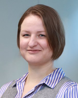 Eine Person mit kurzen, braunen Haaren und einem grauen Pullover mit blauer gestreifter Bluse, die in einem hellen Raum steht.