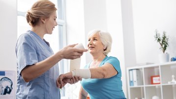 Eine Krankenschwester spricht mit einer älteren Frau, die einen Verband am Arm trägt, in einer hellen, modernen Praxis.