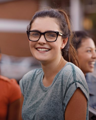Eine Gruppe von vier jungen Erwachsenen steht zusammen, während eine Frau mit Brille und einem lächelnden Gesicht im Vordergrund hervorsticht. Sie trägt ein graues T-Shirt und hat lockiges Haar.
