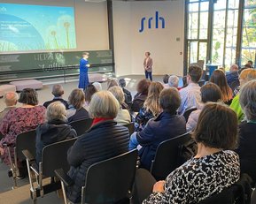 Eine Präsentation in einem großen Raum, mit einem Publikum, das auf zwei Redner auf der Bühne achtet. Ein Bildschirm zeigt Informationen.