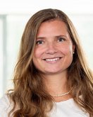 Eine Frau mit langen, lockigen Haaren und einem freundlichen Lächeln trägt ein graues und weißes Oberteil. Sie steht vor einem hellen Hintergrund mit unscharfen Fenstern.