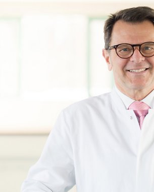 Ein Arzt in einem weißen Kittel mit einer rosa Krawatte steht in einem hellen, modernen Raum und lächelt freundlich.