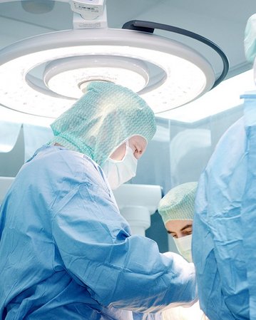Drei Chirurgen in OP-Kleidung führen einen chirurgischen Eingriff unter einem Operationslicht durch. Sie tragen Masken und Hauben, während sie sich auf den Patienten konzentrieren.